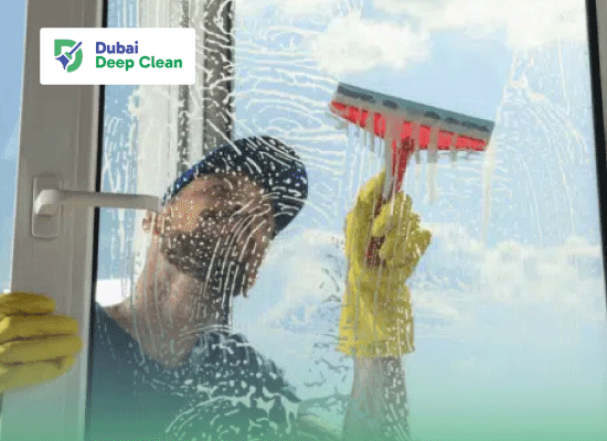 A professional cleaner in a cap and yellow gloves using a squeegee to clean soapy residue from a large balcony glass door under a blue sky.