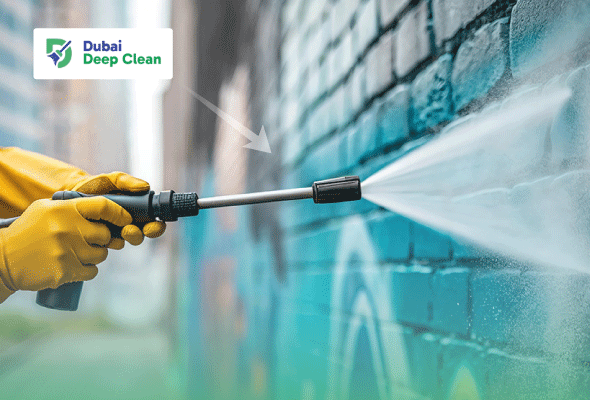 A cleaning professional in yellow protective gloves using a high-pressure water jet to deep clean a brick wall in a garage or outdoor area.
