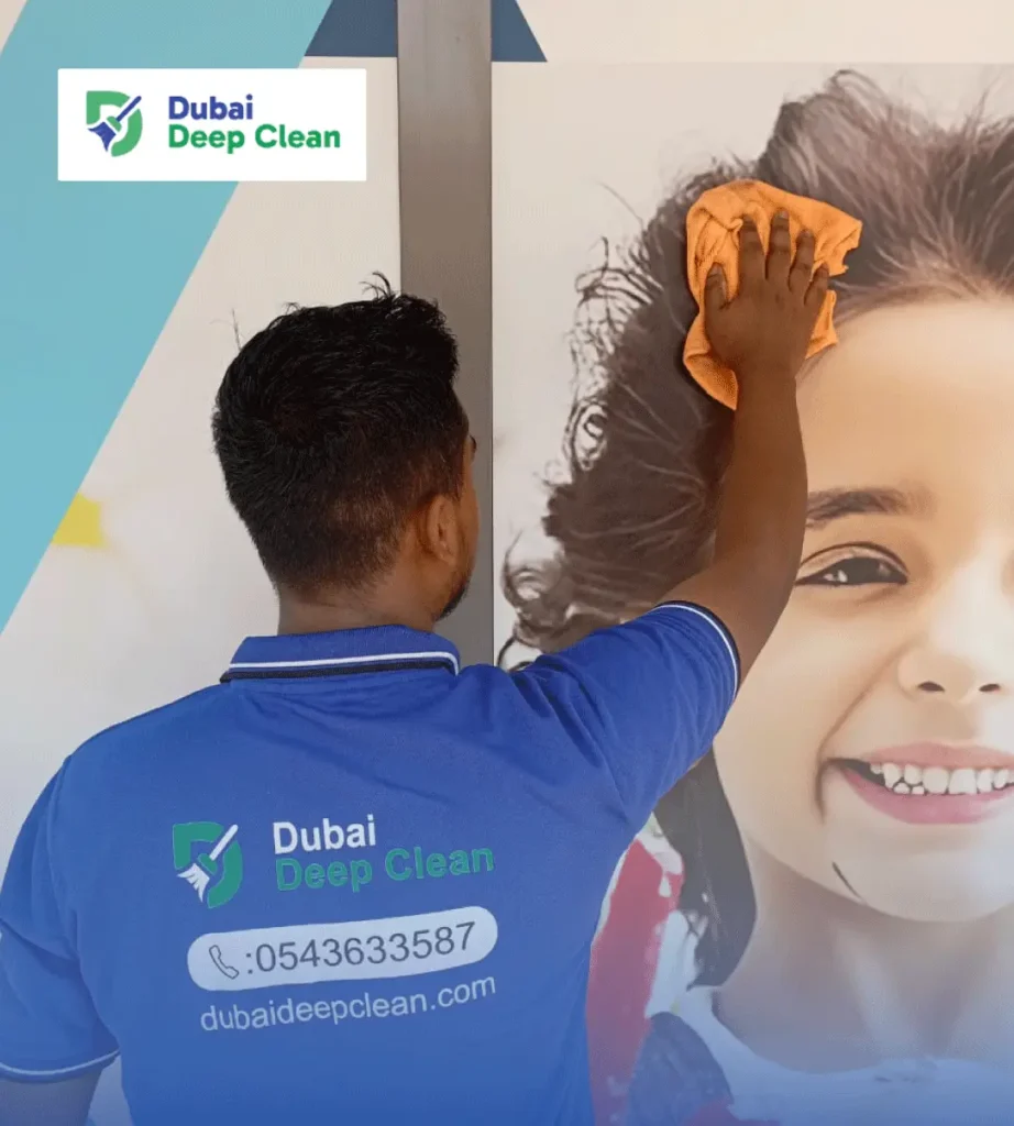 A professional cleaner from Dubai Deep Clean, wearing a blue branded polo shirt, using an orange microfiber cloth to carefully clean a large wall-mounted portrait.