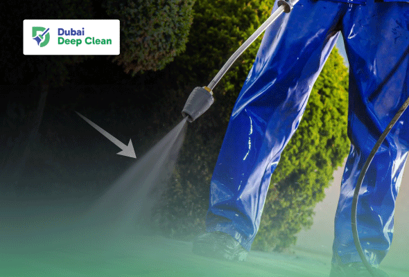 A professional cleaner in blue waterproof protective gear using a high-pressure power washer to clean a stone walkway or outdoor area.