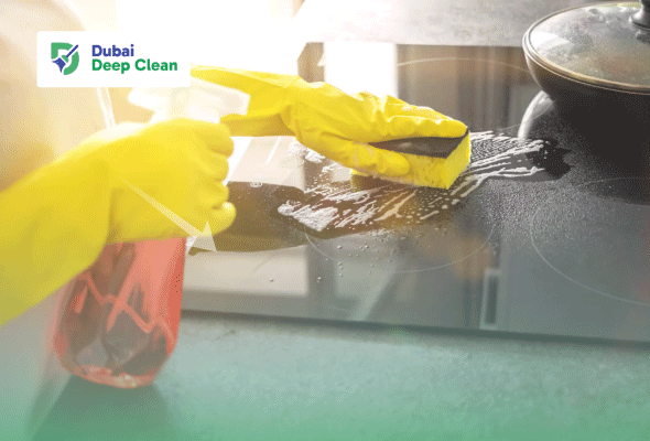 A cleaning expert wearing yellow industrial gloves using a disinfectant spray and a scrubbing sponge to deep clean a greasy black induction stovetop in a professional restaurant kitchen.
